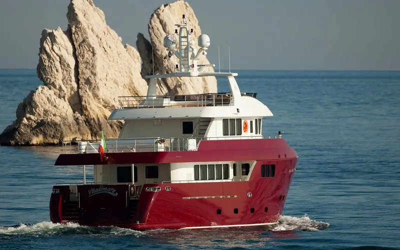 The Darwin Class 86 Vitadimare passes a rock stack on the Italian coast, her red hull and raised pilothouse characteristic of Cantiere delle Marche's explorer line.
