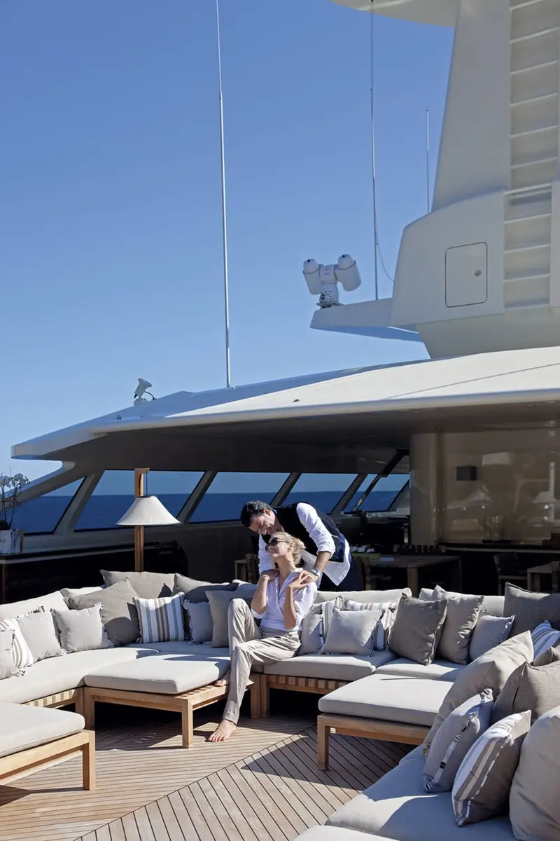 Guests and crew relax on the aft deck beneath a fixed shade structure, drinks laid out on a low table.