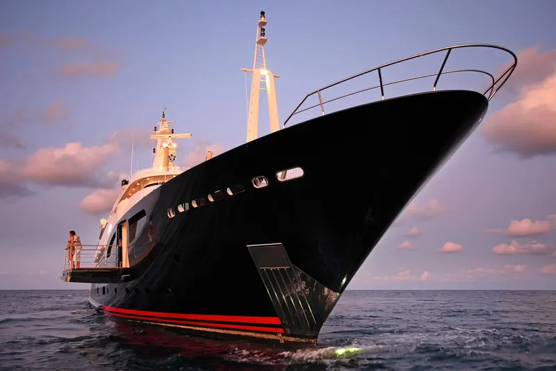 The same dark hull photographed bow-on at dusk, a couple standing on the starboard balcony as the anchor chain runs down into calm water.