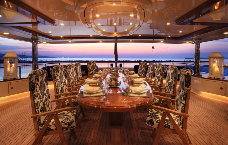 A formal dining room on board with a long table set for a full crew, patterned upholstered chairs and a view out to the sunset over the water.