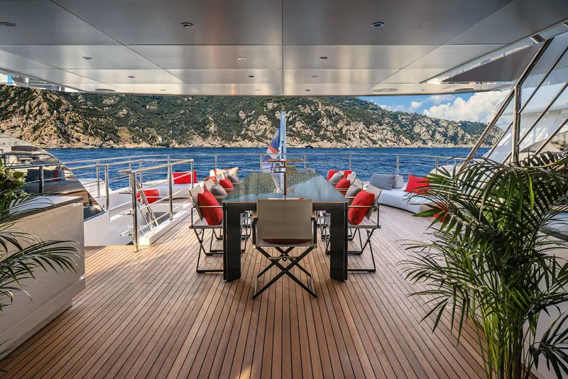 An al fresco dining table on the aft deck with red cushioned chairs and a potted palm for shade.