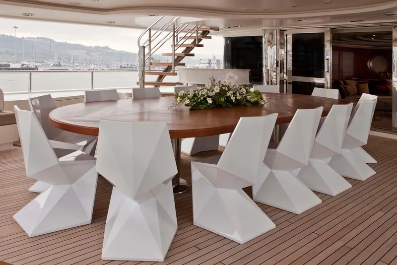 An al fresco dining table on the aft deck surrounded by angular, faceted white chairs, a design detail distinct from CRN's usual upholstered deck furniture.