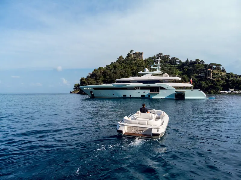 Latona at anchor in Portofino's harbour with a tender approaching her swim platform, the village's coloured houses visible on the far shore.