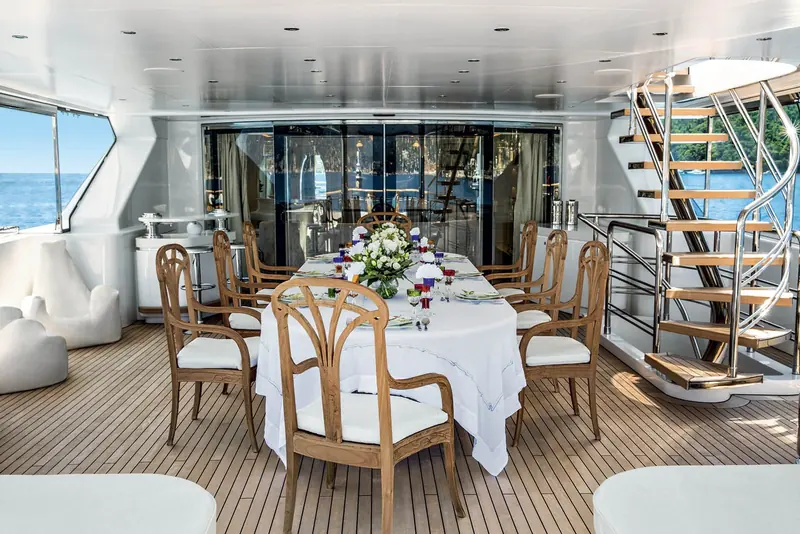 A formal dining room on board with a curved staircase behind the table, white upholstered chairs and a wall of glass looking out to the water.