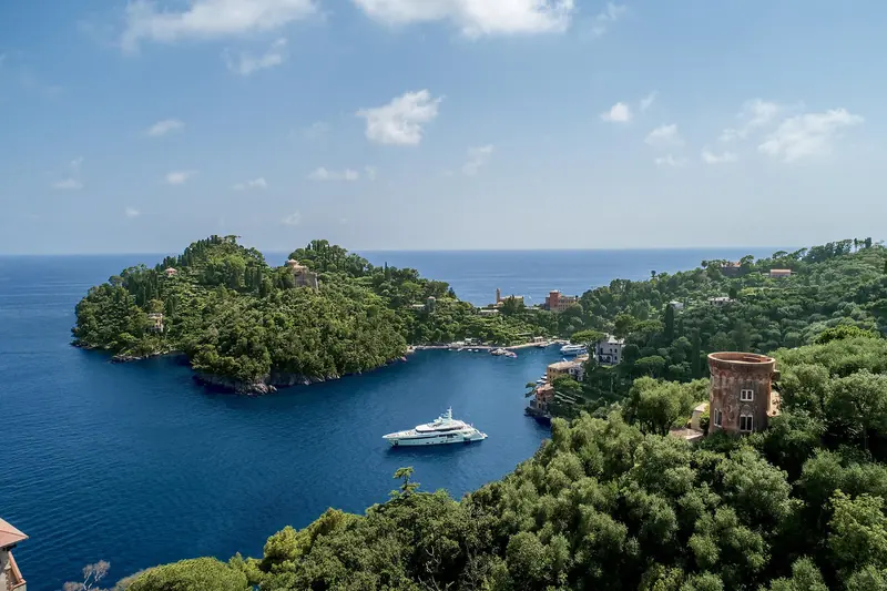 An aerial view of Portofino's promontory and harbour mouth with Latona anchored in the bay below, the medieval watchtower visible on the headland.