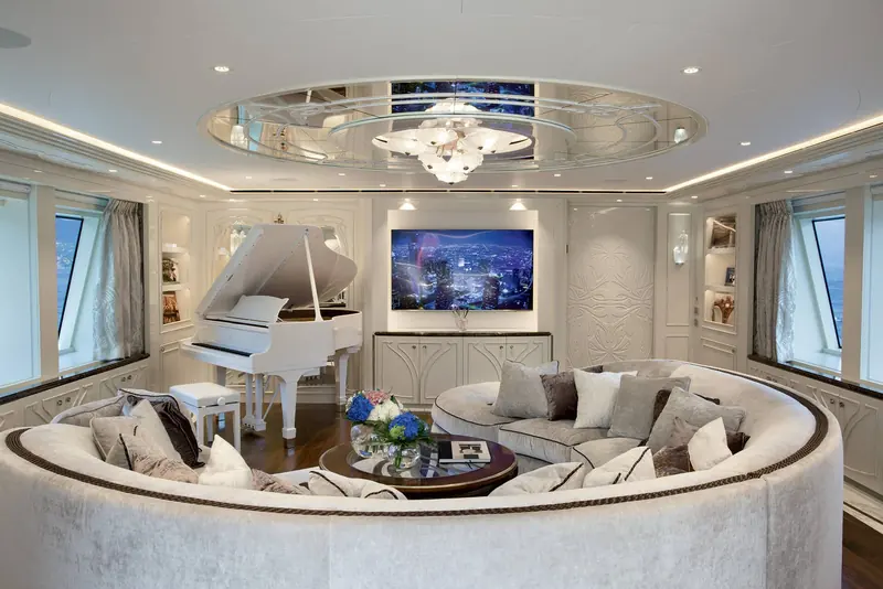 A curved sitting room interior with a white grand piano, built-in bench seating and a wall-mounted screen, photographed in soft daylight.