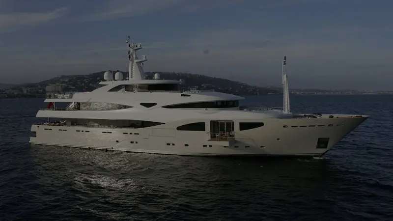 Maraya underway along the French Riviera, a wooded hillside and coastal town visible behind her pale grey hull.