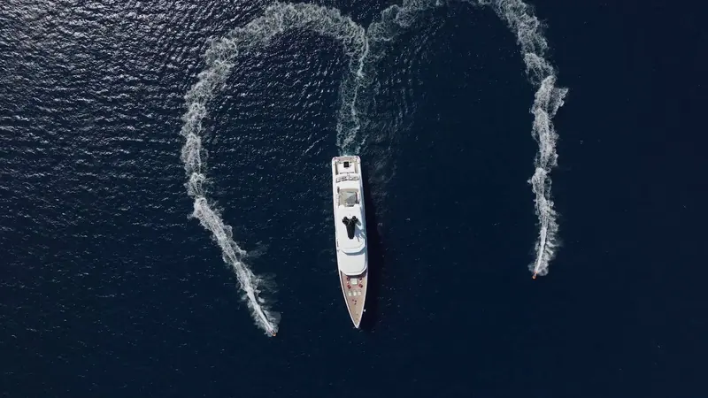 A tight heart-shaped wake traced by Rio's tender turning circles beside the stationary yacht, shot from directly overhead.