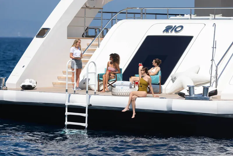 Guests on the transom swim platform beside the painted RIO nameplate, the platform lowered to sea level for direct water access.