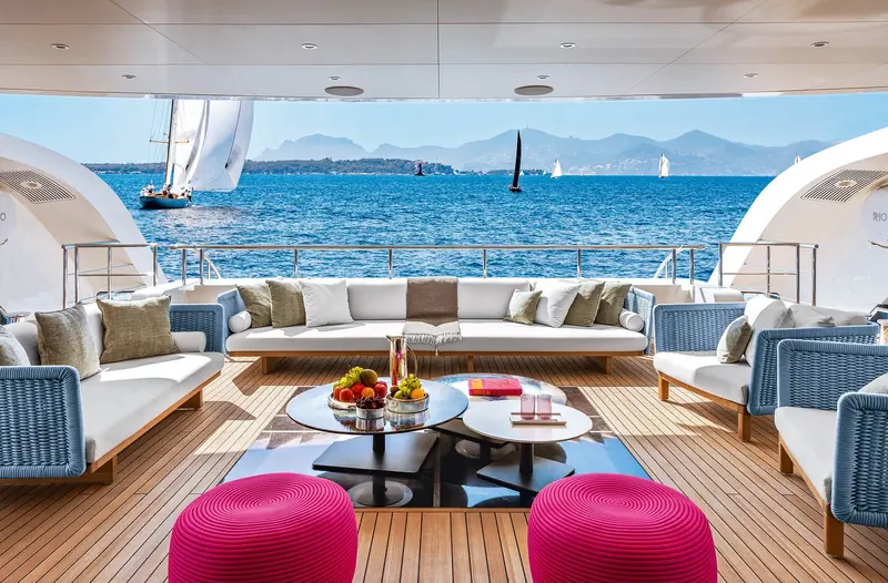 An aft deck lounge set with white sofas, magenta poufs and a round glass table, sailboats visible racing in the bay behind.