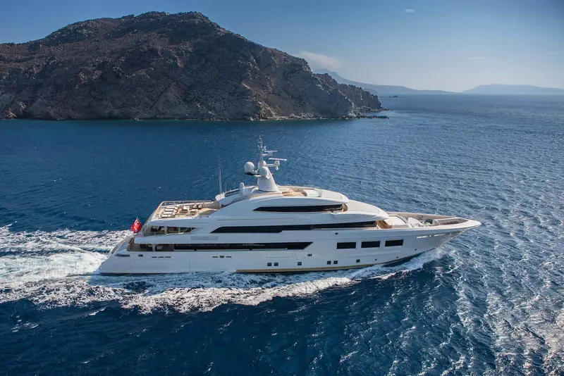 Saramour underway in the Aegean, a bare rocky island rising behind her and the sea deep blue under a clear sky.