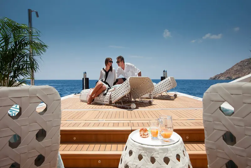An aft-deck lounge area with woven basket-weave furniture and a potted palm, a couple seated in conversation.