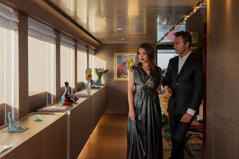 A couple in formal evening wear walk through an interior corridor hung with framed artwork, styled as a lifestyle rather than a specification shot.