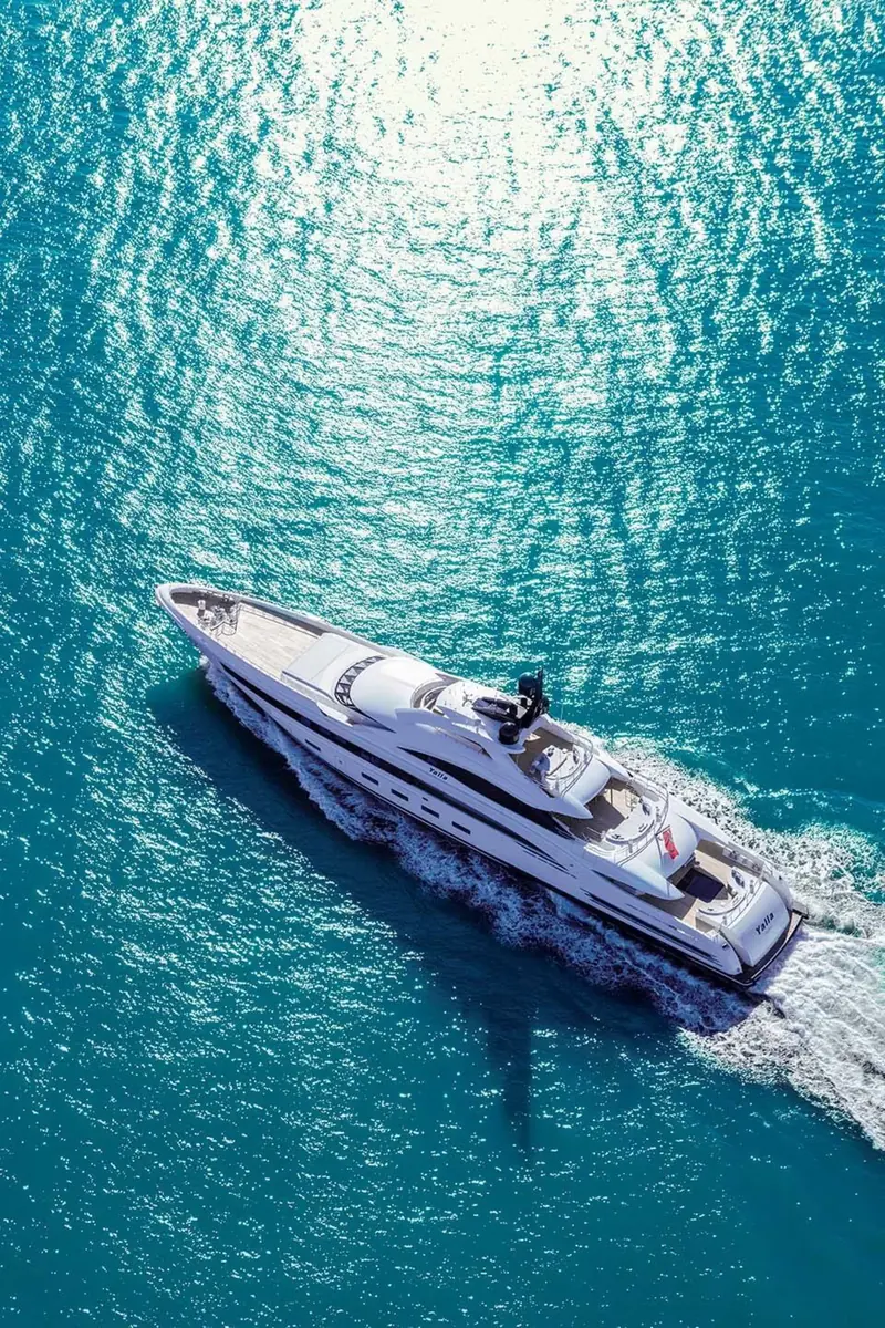 An aerial bow-quarter view of Yalla underway, sunlight scattering across the water's surface behind her.