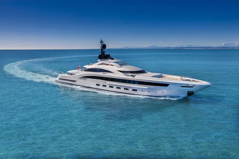 An aft-deck view with a couple relaxing on deck furniture, the yacht's own wake visible astern.