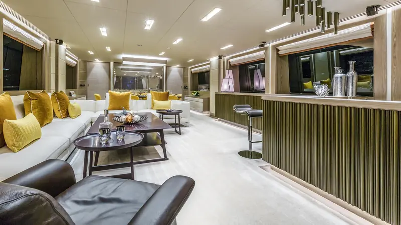 The main deck salon, finished in pale oak with a full-beam bar aft and mustard-yellow accent seating.