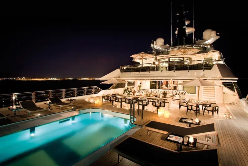 The aft deck lit for the evening, pool glowing turquoise beside a long dining and lounge setup.