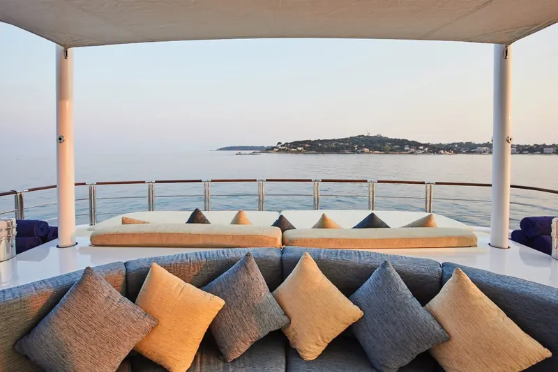The shaded aft lounge, cushions set out under an awning with a headland visible astern.