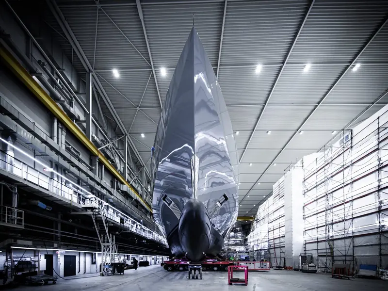 The mirror-polished hull inside the covered shed before paint, dwarfing the scaffolding either side.