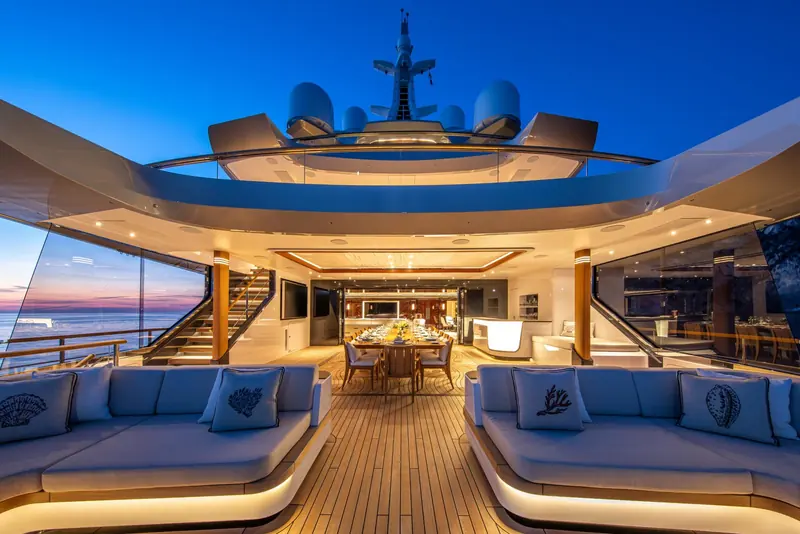 The main deck saloon opens straight onto the aft deck lounge through full-height glass, so the dining table and the outdoor sofas read as one room.