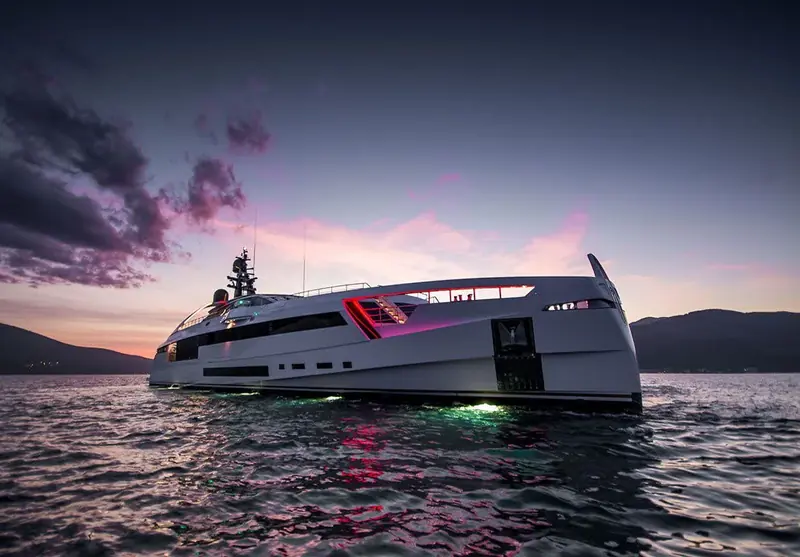 Aurora at anchor in Kotor Bay under a violet dusk sky, her hull-side lighting glowing green and pink in the water.