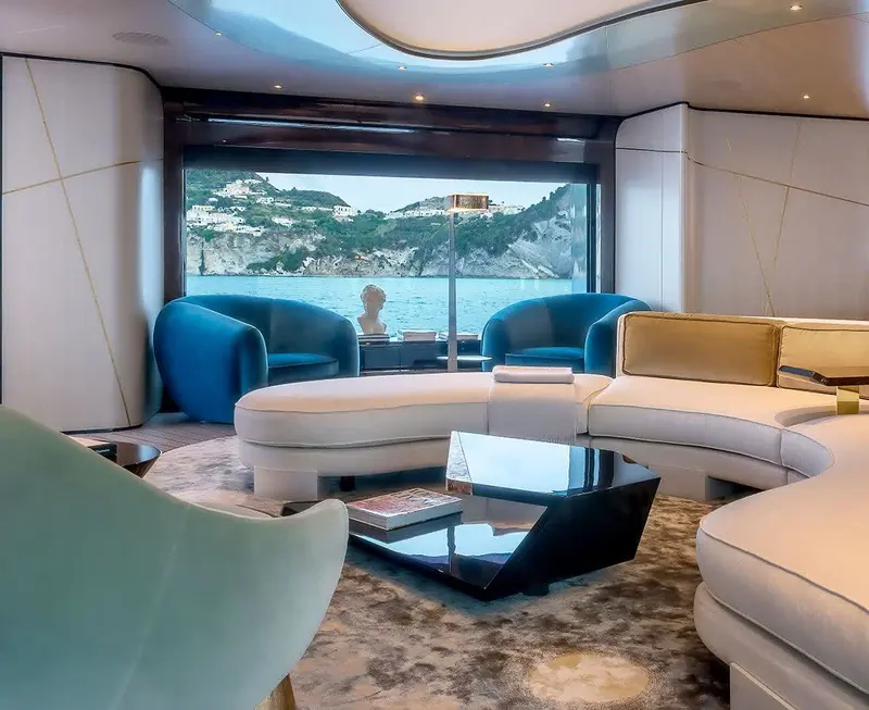 The main saloon's curved seating in duck-egg blue, with a full-height window framing the anchorage outside.