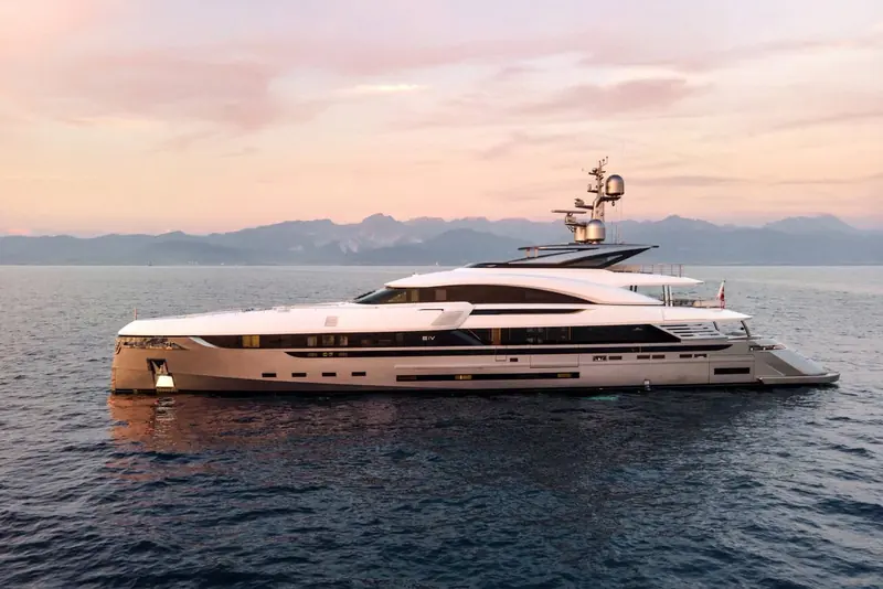 At anchor off a mountainous coastline at sunset, EIV's name spelled out in dark glass along the hull side.
