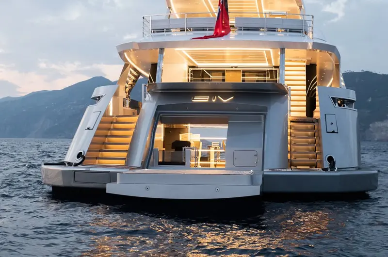 The stern at dusk, twin staircases and a fold-out beach club lit warm against the darkening hillside behind.