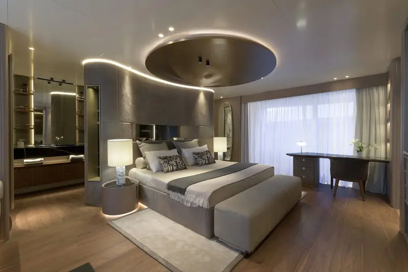 A guest cabin finished in grey oak with a large oval mirror set into a backlit ceiling recess above the bed, and a full en-suite bathroom visible through the open door.
