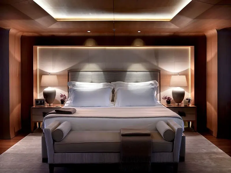 The master suite, finished in dark wood veneer with a symmetrical pair of nightstand lamps and a bench at the foot of the bed, in the same warm register as the main salon.