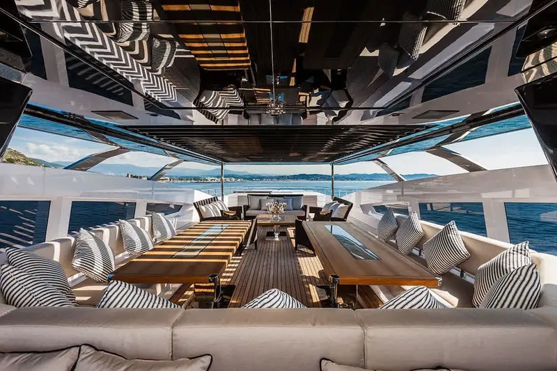 Twin teak dining tables under a retractable hardtop, its underside mirrored so the striped cushions repeat overhead.