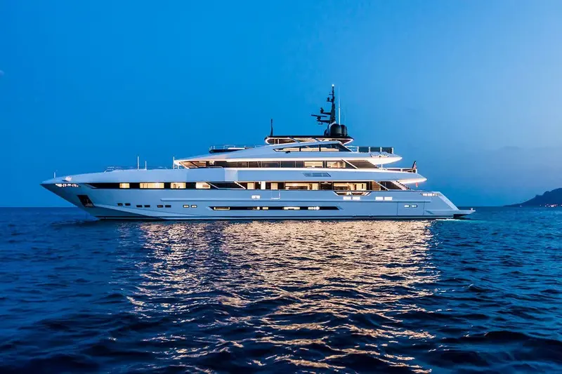 Broadside at dusk, deck lights coming on as she holds position off a lit shoreline.