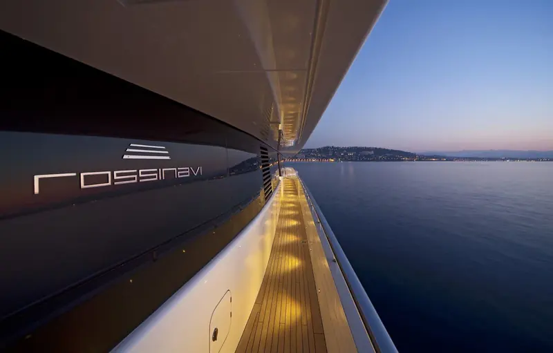 A side-deck detail at dusk, with the Rossinavi nameplate lit against the hull and the coast glowing in the distance, the walkway lighting designed for guests moving forward after dark.