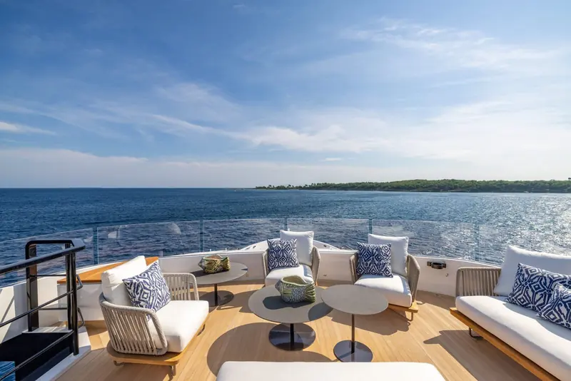 Aft deck lounge at anchor off a wooded coastline, cushions in navy and white around a low central table.