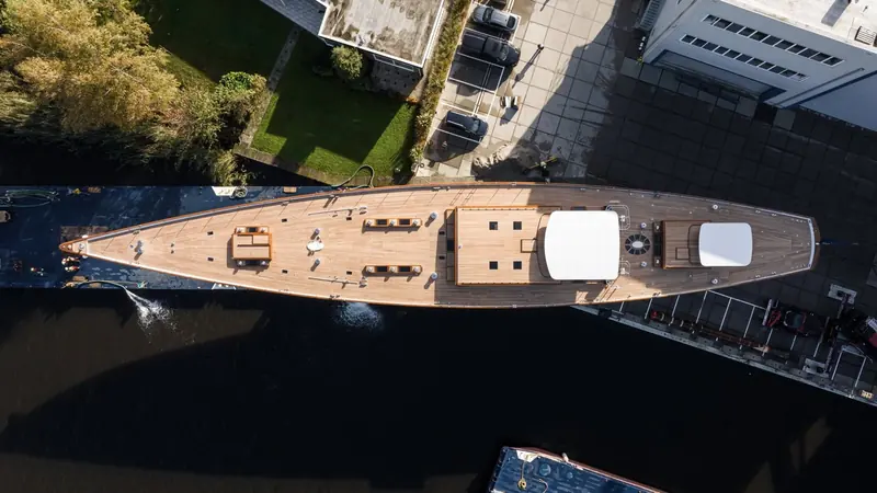 A straight-down aerial shows the full deck plan: teak decks running unbroken from bow to the aft cockpit, with the two masts stepped well forward of the deckhouse.