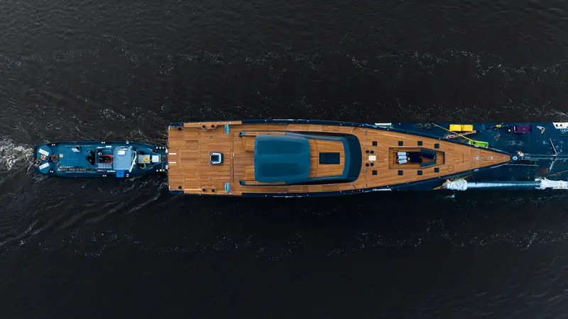 Fresh off the build hall and under tow, the teak foredeck still bare of running rigging and the yard's tools still lying where the fit-out crew left them.