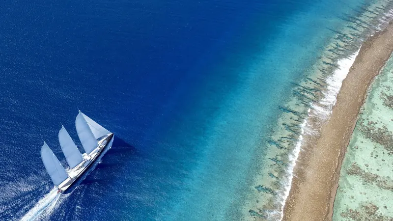 A straight-down aerial over the reef pass shows the three-masted schooner rig at full spread, wake curling flat off a hull built for long ocean passages, not just harbour showing-off.