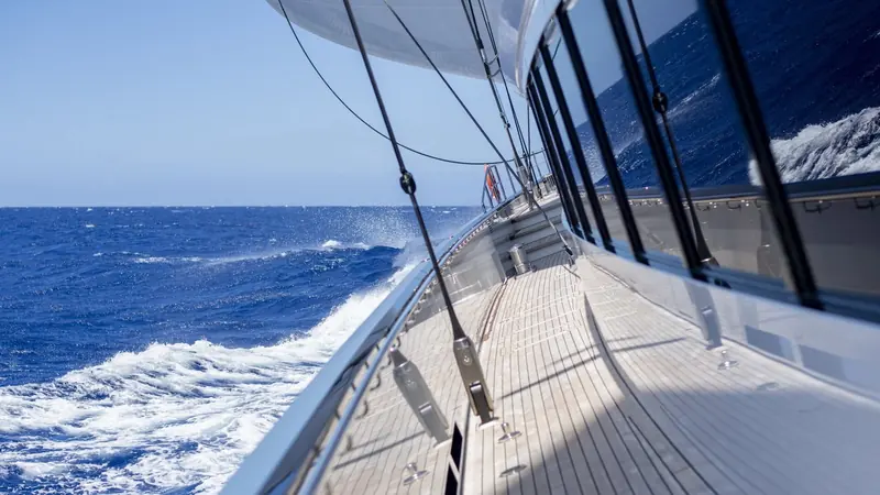 Spray breaking against the topsides at speed - the teak side deck and mirror-polished stanchions are the only clue this is a superyacht rather than a racing sloop.