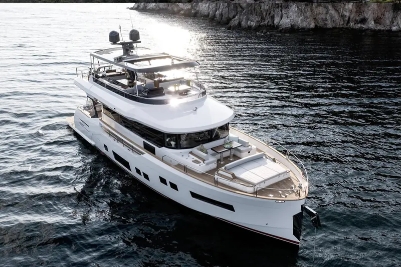 Cruising close along a rocky shoreline in bright sun, flybridge sunpads and helm seating visible under the open hardtop.