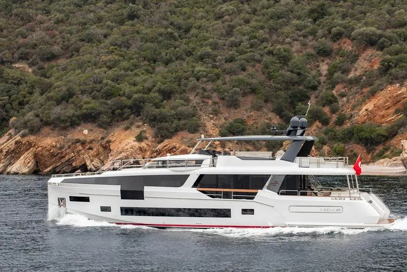 Passing close under a pine-topped headland, the red boot-stripe and dark hull windows visible along the full main-deck run.
