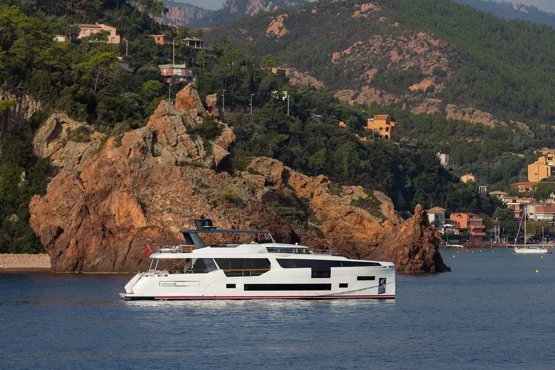 At anchor in a quiet cove with hillside villas behind her, giving a sense of scale against the coastline she is built to cruise.
