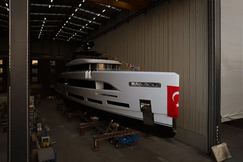 The bow carries a Turkish flag for the traditional launch blessing as the hull clears the shed doors, signal flags strung from the mast to the roofline.