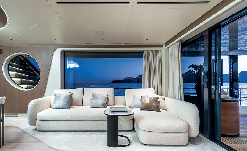 A lounge sofa aboard an Arcadia A80, with the water visible through the saloon window at dusk.