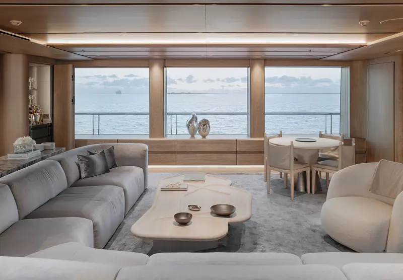 An aft lounge and bar on Entourage, with an organic-edge coffee table and a coastal view through full-height glazing.