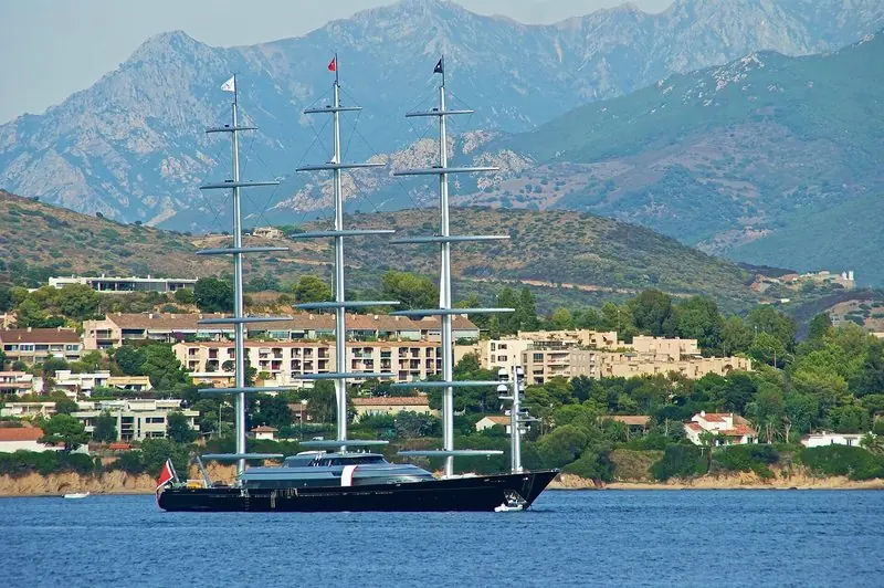 Three unstayed masts mark Maltese Falcon at anchor off a steep, built up shore.