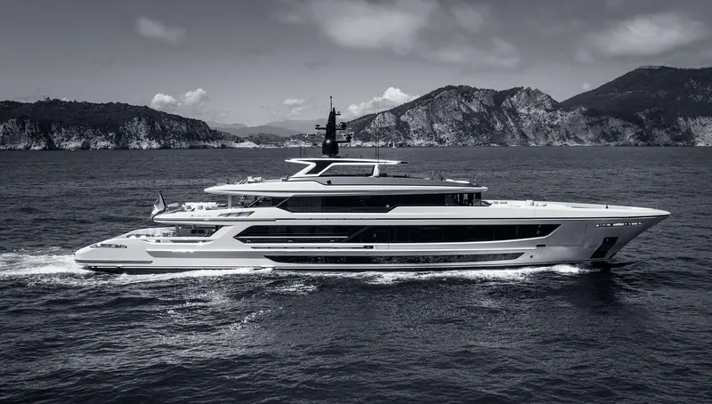 A white superyacht runs close along a rocky headland in flat water, three decks stepped back from an almost plumb bow and a long unbroken band of main deck glass down her side.