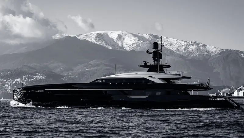 The dark hulled superyacht Geosand runs off a coastal city with snow covered mountains rising behind her, her name in pale lettering on the superstructure side.