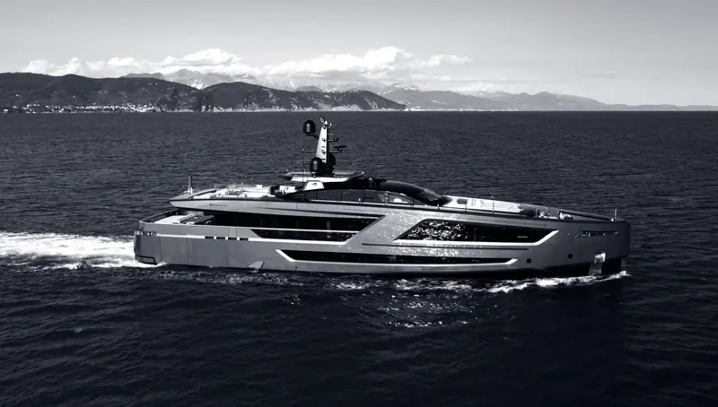 A grey hulled fast motor yacht crosses flat water off a mountainous coast, the Baglietto name and emblem set on the bulwark above her main deck windows.