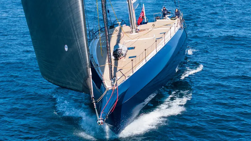 One of the practice's performance sloops drives bow-first into a wave, two crew at the helm under a red ensign as spray bursts off the bow.