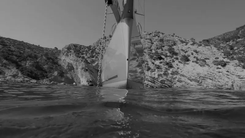 Low to the waterline, the bow of a yacht at anchor rises against a rocky shore.